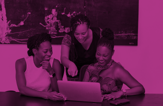 The photo depicts three black women looking at a laptop screen and smiling. Two of them are sitting at a desk, a third one is standing in the middle and pointing at the screen.